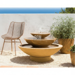 A low-profile modern 3-Tier Cascading Bowl Fountain made of natural earth-toned stone, featuring wide, shallow basins spilling water, placed in a sunny Zen garden setting with a modern wire chair.